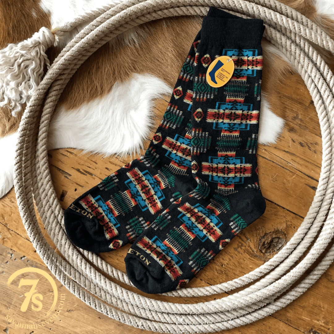 A pair of Chief Joseph Pendleton Crew Socks by Savannah Sevens western life{&}style with colorful geometric patterns and a yellow tag rests inside a coiled rope on a wooden floor, with part of a fur rug visible in the background.