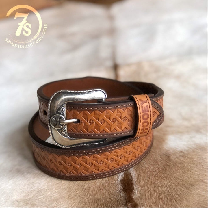 The Dayton Belt {mens} by Savannah Sevens western life{&}style is a tooled basketweave leather belt with a decorative silver buckle, stamped with HOOT near the end, coiled on cowhide. Logo appears in the top left corner.