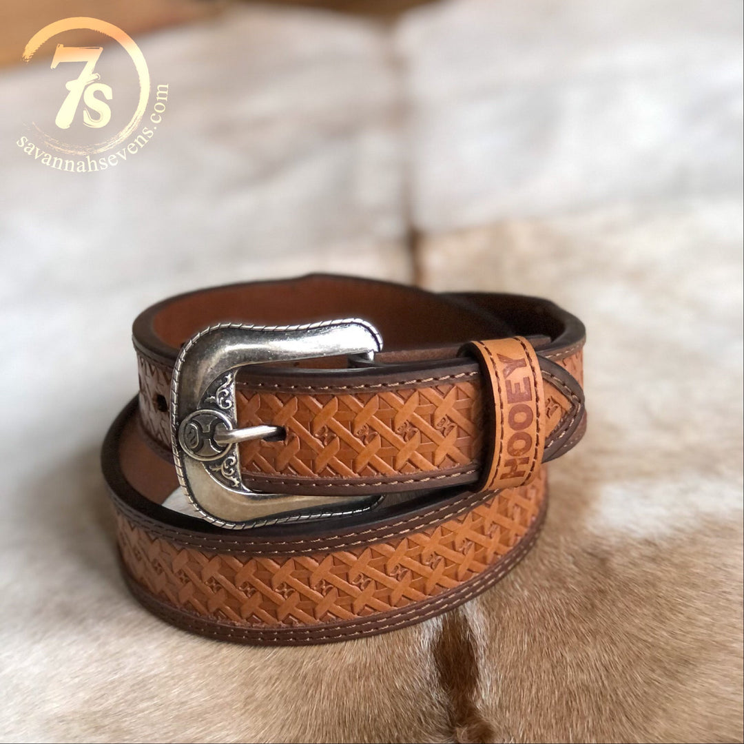 The Dayton Belt {mens} by Savannah Sevens western life{&}style is a tooled basketweave leather belt with a decorative silver buckle, stamped with HOOT near the end, coiled on cowhide. Logo appears in the top left corner.