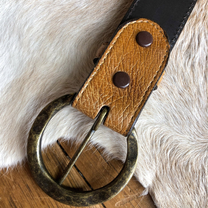 A close-up of the Du Bois Belt by Savannah Sevens western life{&}style, featuring a saddle tan textured brown strap, round brass buckle, and two metal rivets, resting on animal hide and wood.