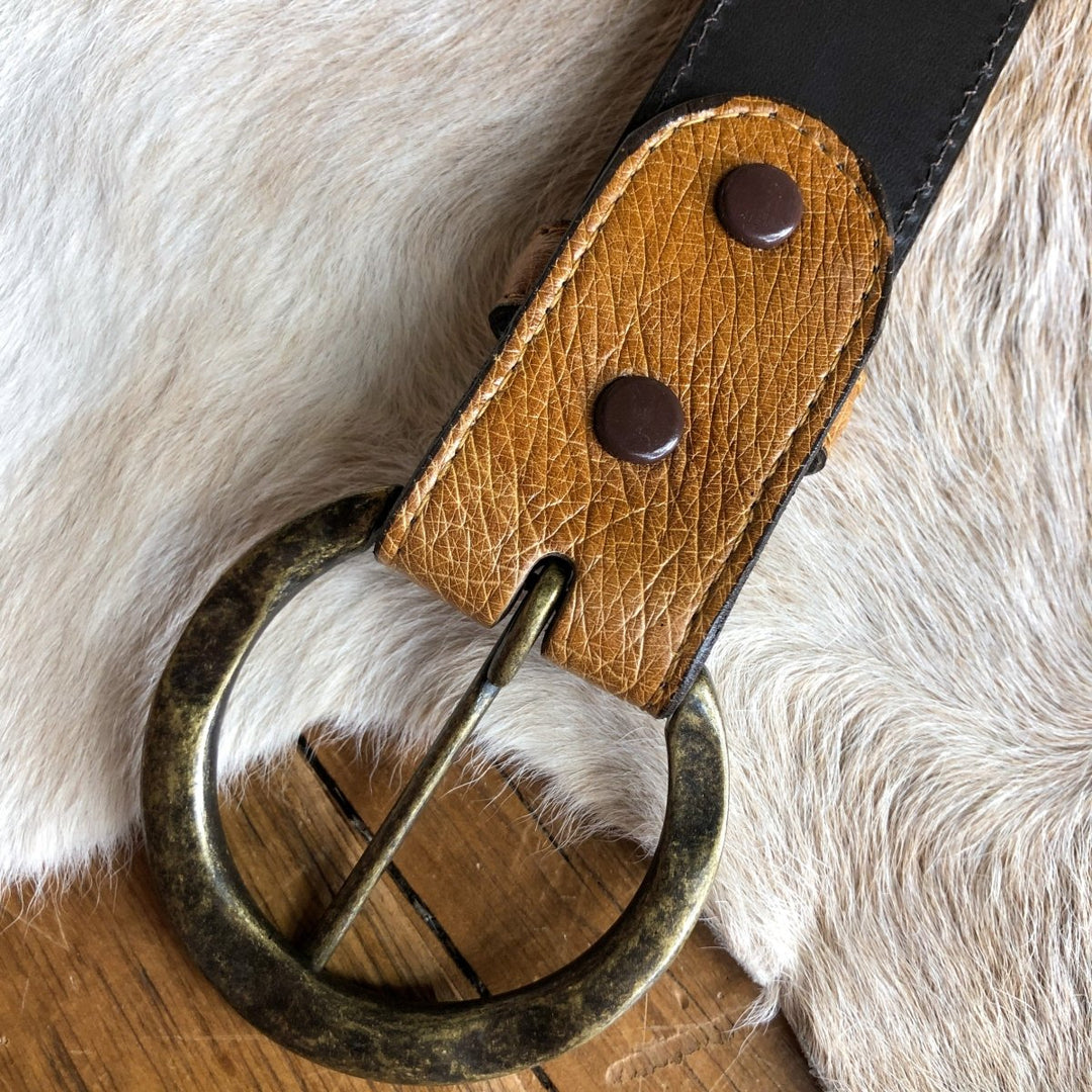 A close-up of the Du Bois Belt by Savannah Sevens western life{&}style, featuring a saddle tan textured brown strap, round brass buckle, and two metal rivets, resting on animal hide and wood.