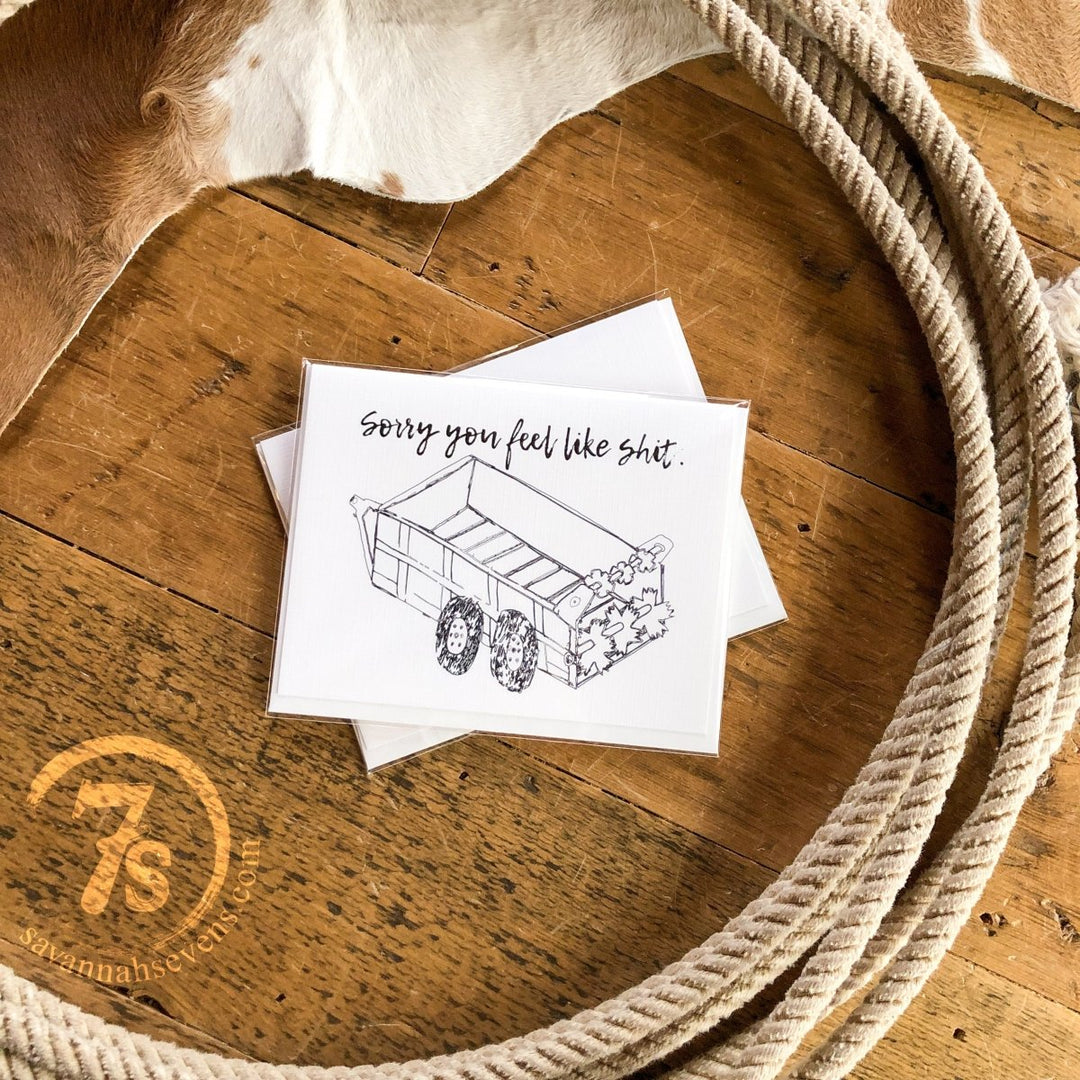 The Hungry Canyon Feel Better Card, featuring a dump truck, sits on rustic wood with rope and cowhide in the background—an encouragement card offering support during tough times.