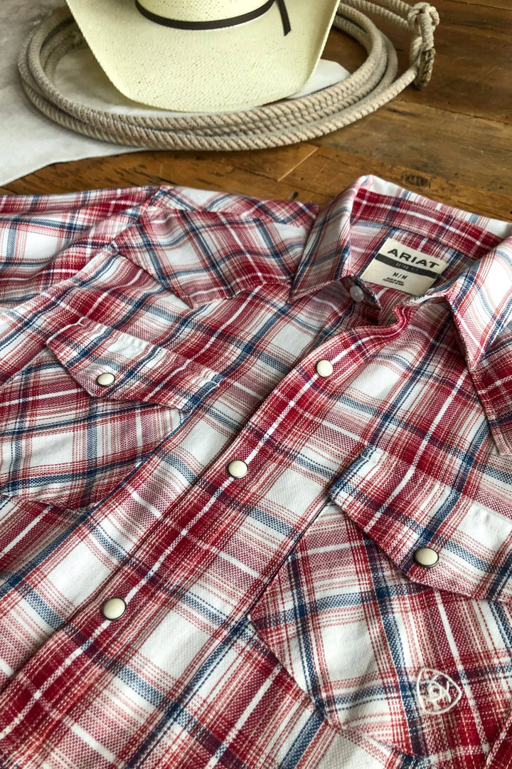 The Glenwood Ariat Men's Plaid Button Up from Savannah Sevens western life{&}style, in red, white, and blue with snap buttons, is displayed on a wooden surface with a white cowboy hat in the background.