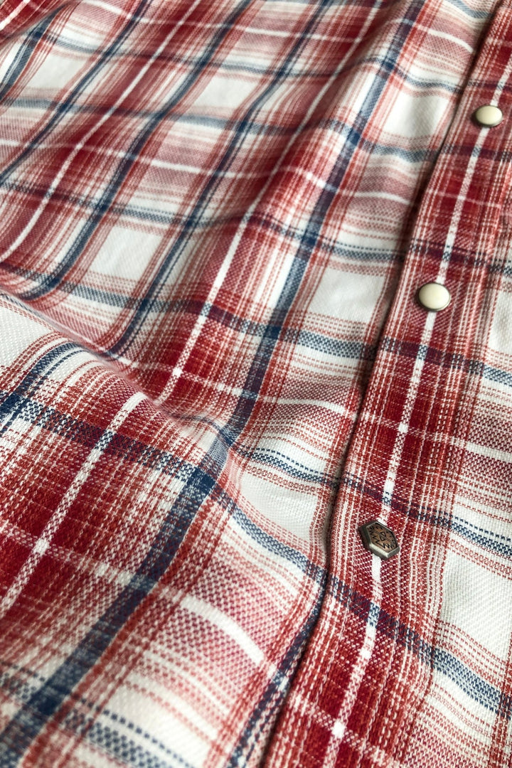 A close-up of the Glenwood Ariat Men's Plaid Button Up by Savannah Sevens western life{&}style in red, white, and blue plaid, with white pearl snap buttons and a detailed woven texture.
