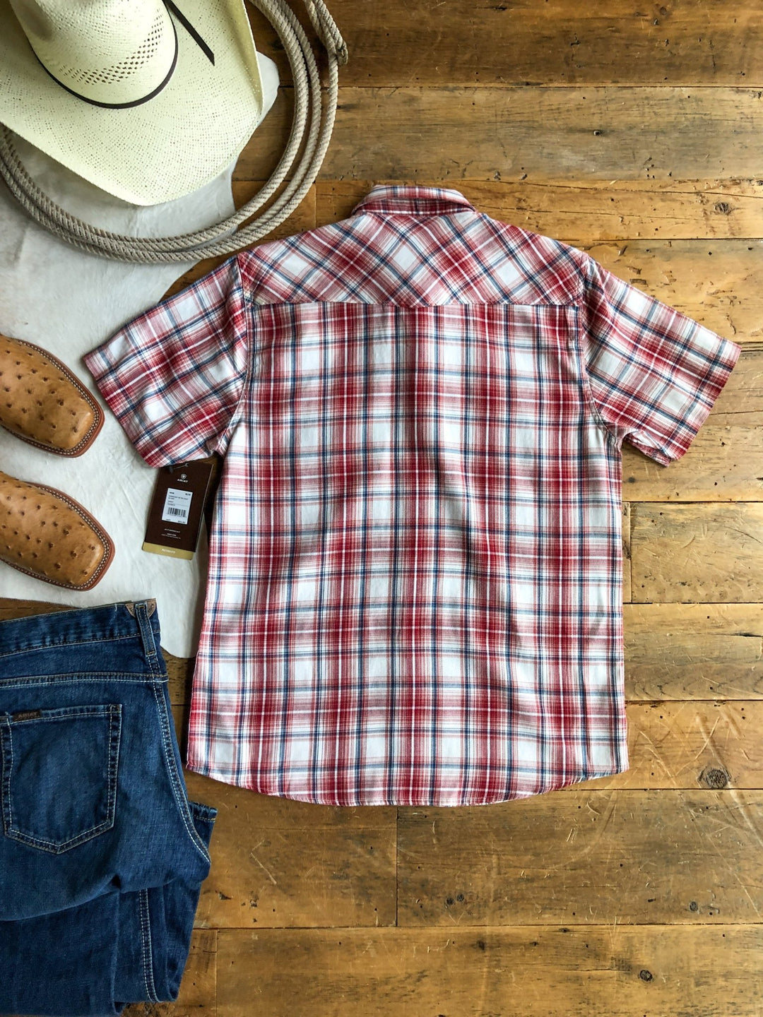 The Glenwood Ariat Men's Plaid Button Up by Savannah Sevens western life{&}style is laid flat, back facing up, on a wooden floor with blue jeans, brown shoes, a coiled rope, and a white cowboy hat nearby.