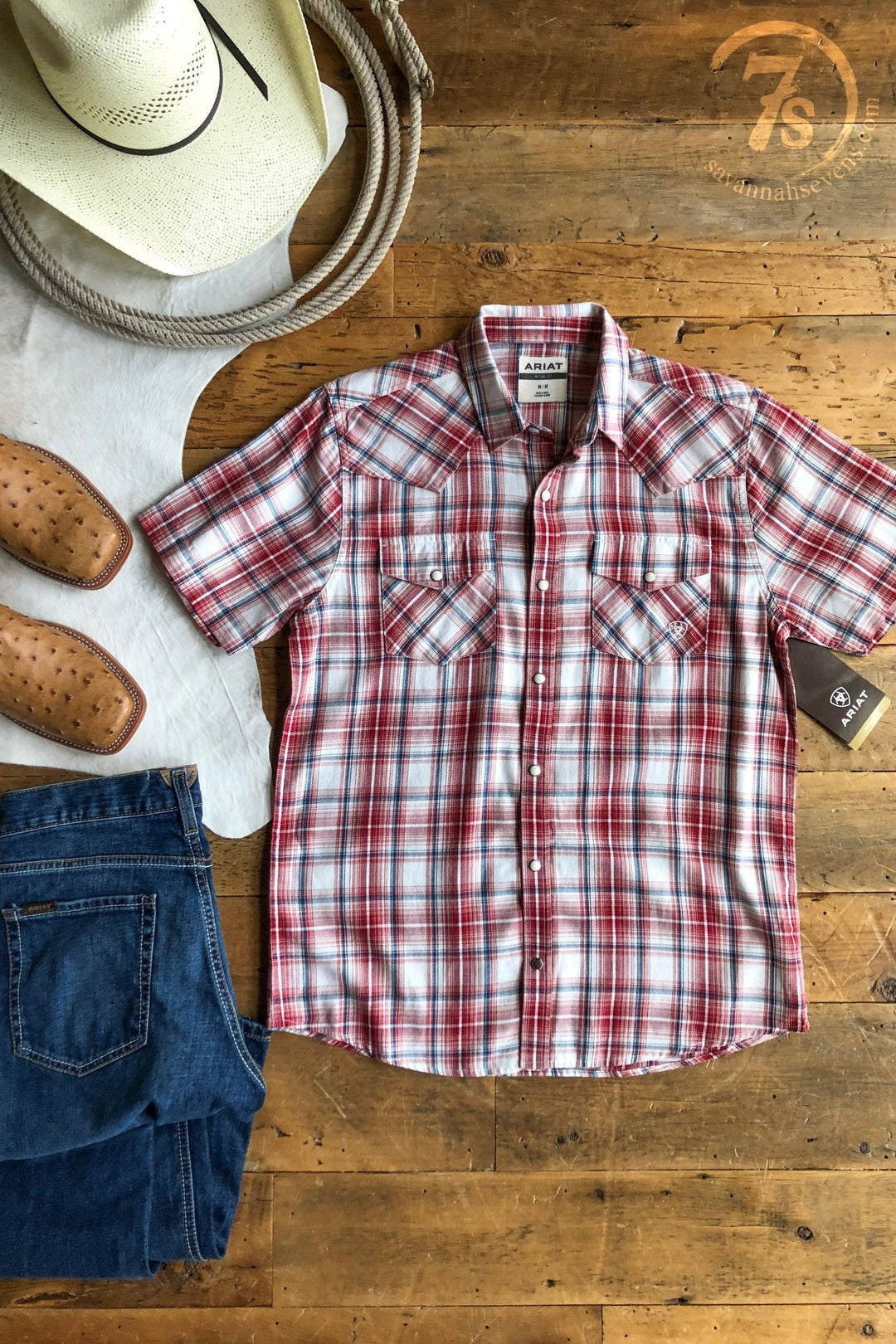 The Glenwood Ariat Men's Plaid Button Up from Savannah Sevens western life{&}style is displayed flat on a wooden floor with jeans, cowboy boots, a white cowboy hat, and a coiled rope arranged around it.