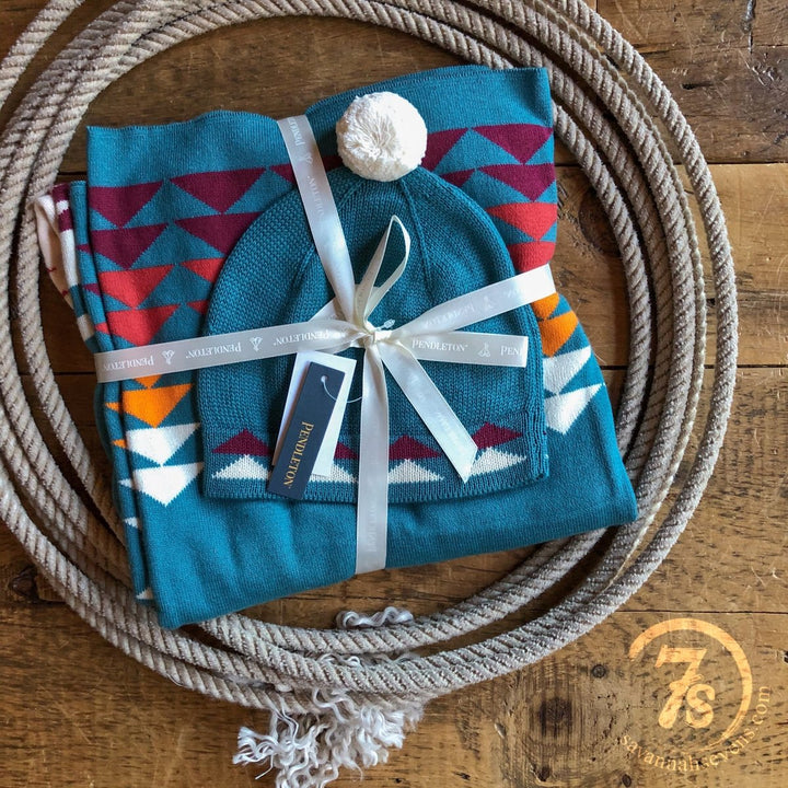 A folded Harding Teal blanket and matching beanie by Savannah Sevens western life{&}style, featuring vibrant southwest patterns, are tied with a white ribbon and rest inside a coiled rope on rustic wood. A circular logo appears in the corner.