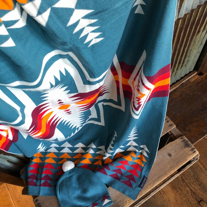 A Harding Teal blanket and matching baby beanie by Savannah Sevens western life&style, featuring white, orange, and red southwest patterns, are draped over a wooden crate; the beanie includes a white pom-pom on top.