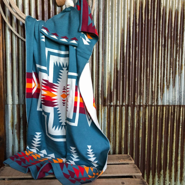 A Harding Teal blanket by Savannah Sevens western life&style, with bold southwest patterns in white, red, orange, and yellow, hangs over a wooden hook against corrugated metal and a rustic crate.