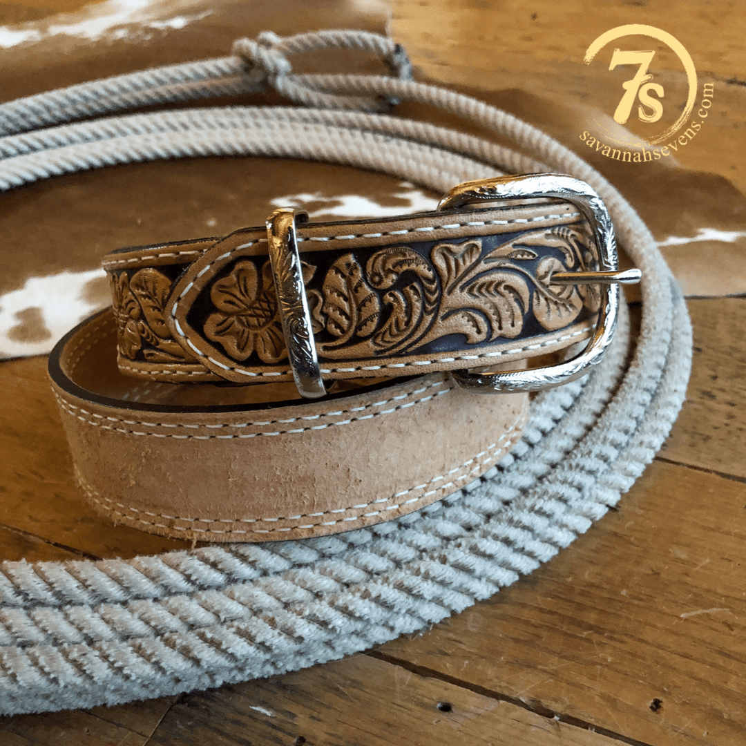 The Hiram Belt from Savannah Sevens western life{&}style, featuring tooled leather with floral designs and a silver buckle, is coiled on wood. A cowhide and the “7s ” logo are seen in the background.