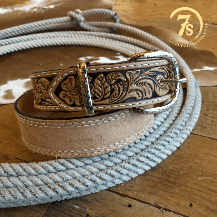 The Hiram Belt from Savannah Sevens western life{&}style, featuring tooled leather with floral designs and a silver buckle, is coiled on wood. A cowhide and the “7s ” logo are seen in the background.