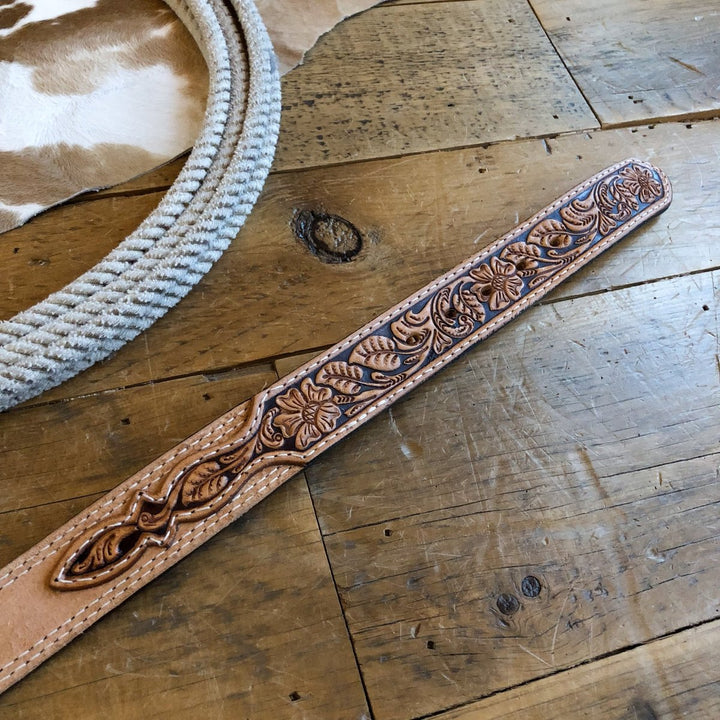 The Hiram Belt from Savannah Sevens western life{&}style, featuring floral tooled leather, lies on a rustic wooden table next to a coiled rope and cowhide.