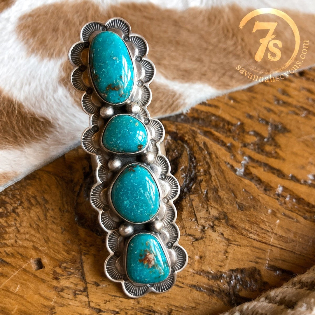 The Keomah Ring by Savannah Sevens western life{&}style features four vertically set turquoise stones of varying sizes in a scalloped sterling silver setting, showcasing Southwest style against a cowhide pattern background on wood.