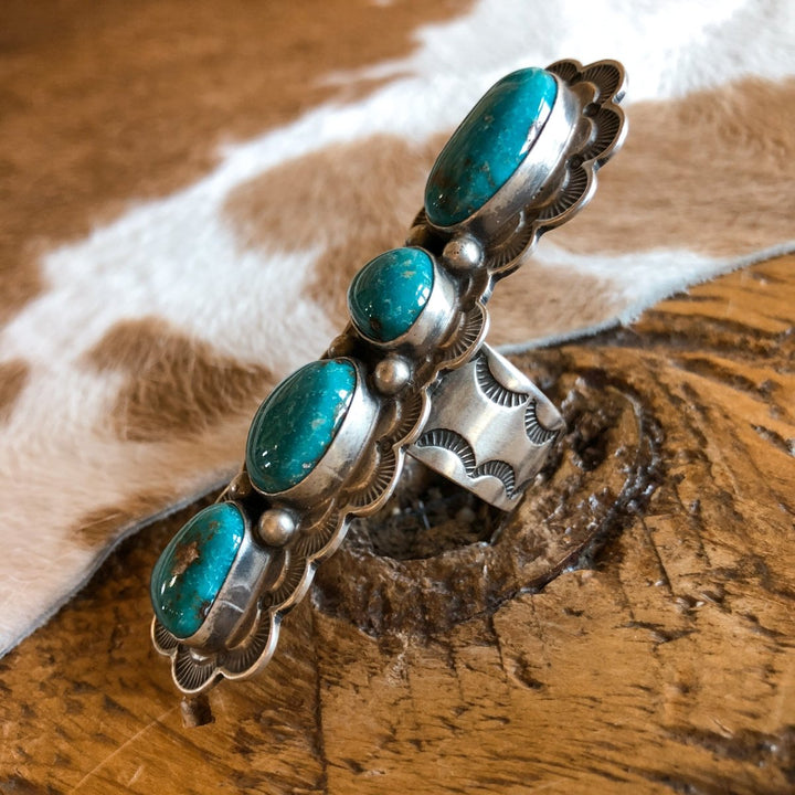 The Keomah Ring by Savannah Sevens western life{&}style features four turquoise stones and engraved sterling silver, displayed on rustic wood with cowhide in the background for a true Southwest jewelry vibe.