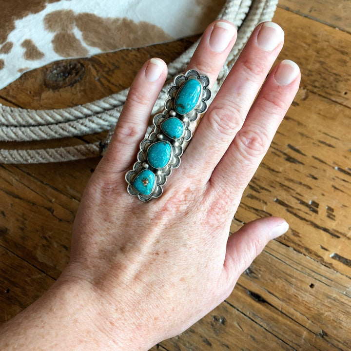 A fair-skinned hand wears the Keomah Ring by Savannah Sevens western life{&}style, a large turquoise piece set in sterling silver, resting on wood beside coiled rope and animal hide.