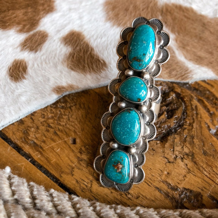 The Keomah Ring by Savannah Sevens western life{&}style features four large turquoise stones set in a sterling silver, ornate scalloped bezel—an eye-catching Southwest jewelry piece displayed on rustic wood.