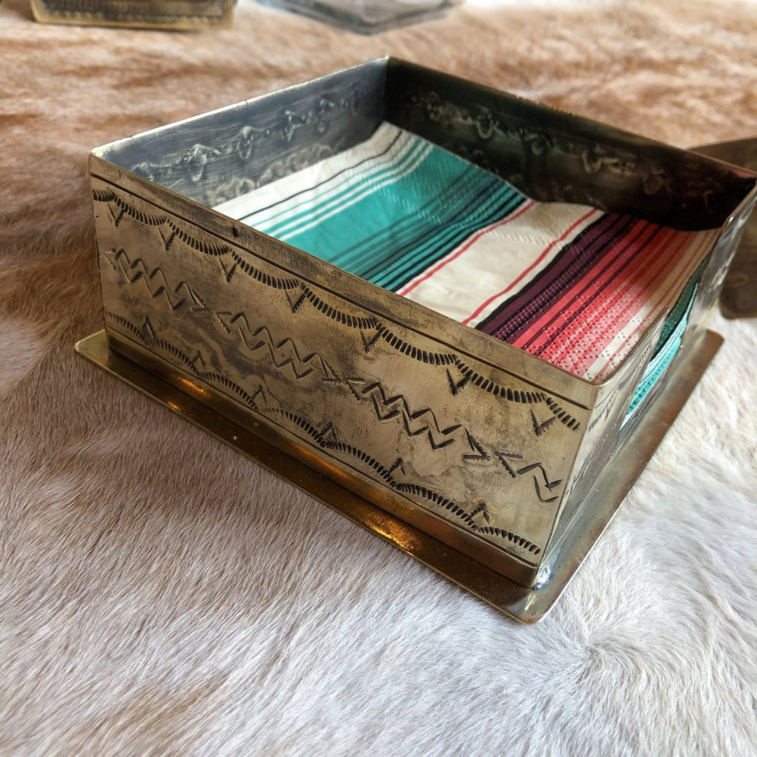 The La Puebla Cocktail Napkin Holder by Savannah Sevens western life{&}style features intricate southwest-inspired engravings on German silver and holds striped red, green, and white napkins. It sits atop a soft, light-colored surface.