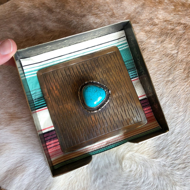 A hand holds the La Puebla Cocktail Napkin Holder by Savannah Sevens western life{&}style, a square metal box with a striped cloth inside and a large silver and turquoise ring on the lid, set against light brown fur for a classic southwest look.