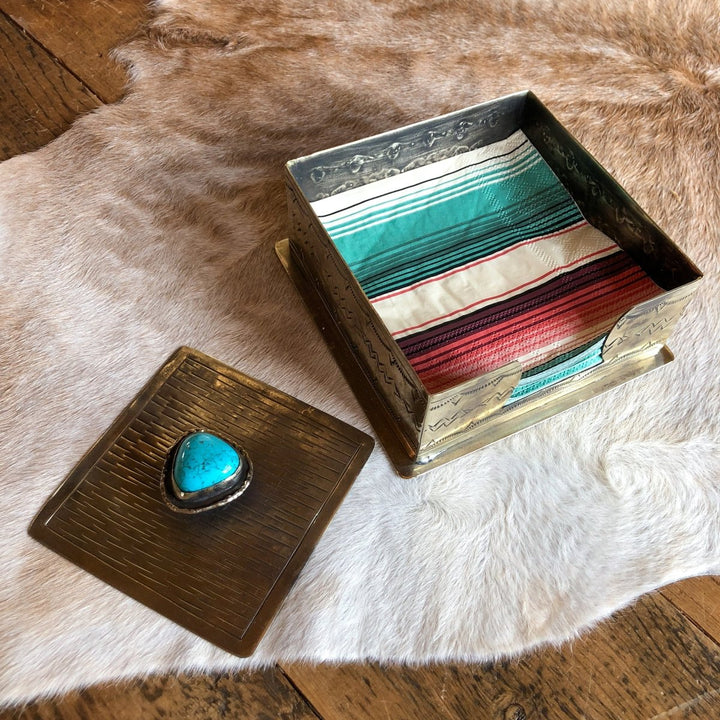 The La Puebla Cocktail Napkin Holder by Savannah Sevens western life{&}style features engraved southwest patterns and a turquoise stone on its lid, displayed open to reveal a colorful striped cloth inside on a wooden floor.