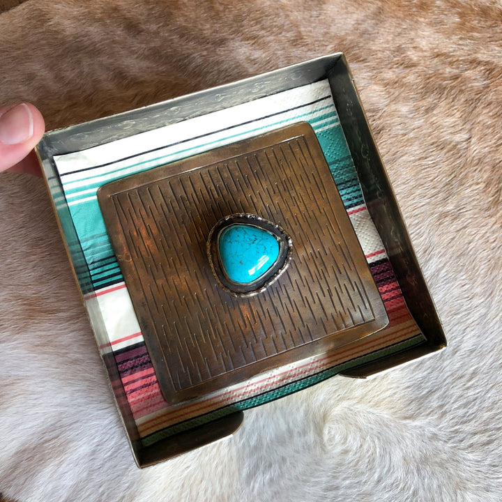 A hand holds the La Puebla Cocktail Napkin Holder by Savannah Sevens western life{&}style, a square metal box with a striped cloth inside and a large silver and turquoise ring on the lid, set against light brown fur for a classic southwest look.