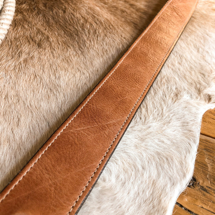 The Langston Belt by Savannah Sevens western life{&}style, featuring tan leather and an interchangeable buckle, rests on light brown and white cowhide with a wooden surface at the edge.