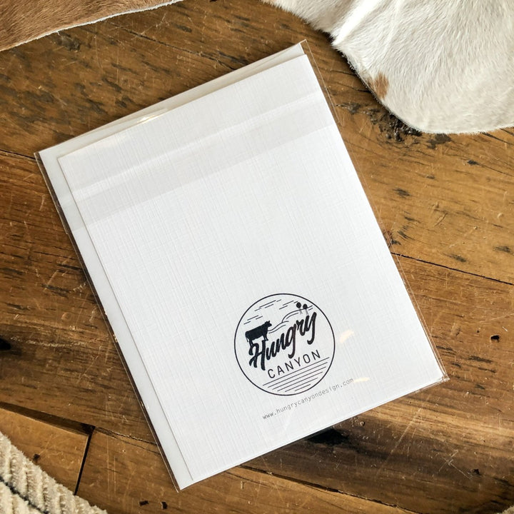 A white "Love You More Than Bacon" card by Hungry Canyon, resembling a blank greeting card, is in clear plastic packaging on a wooden surface next to cowhide.