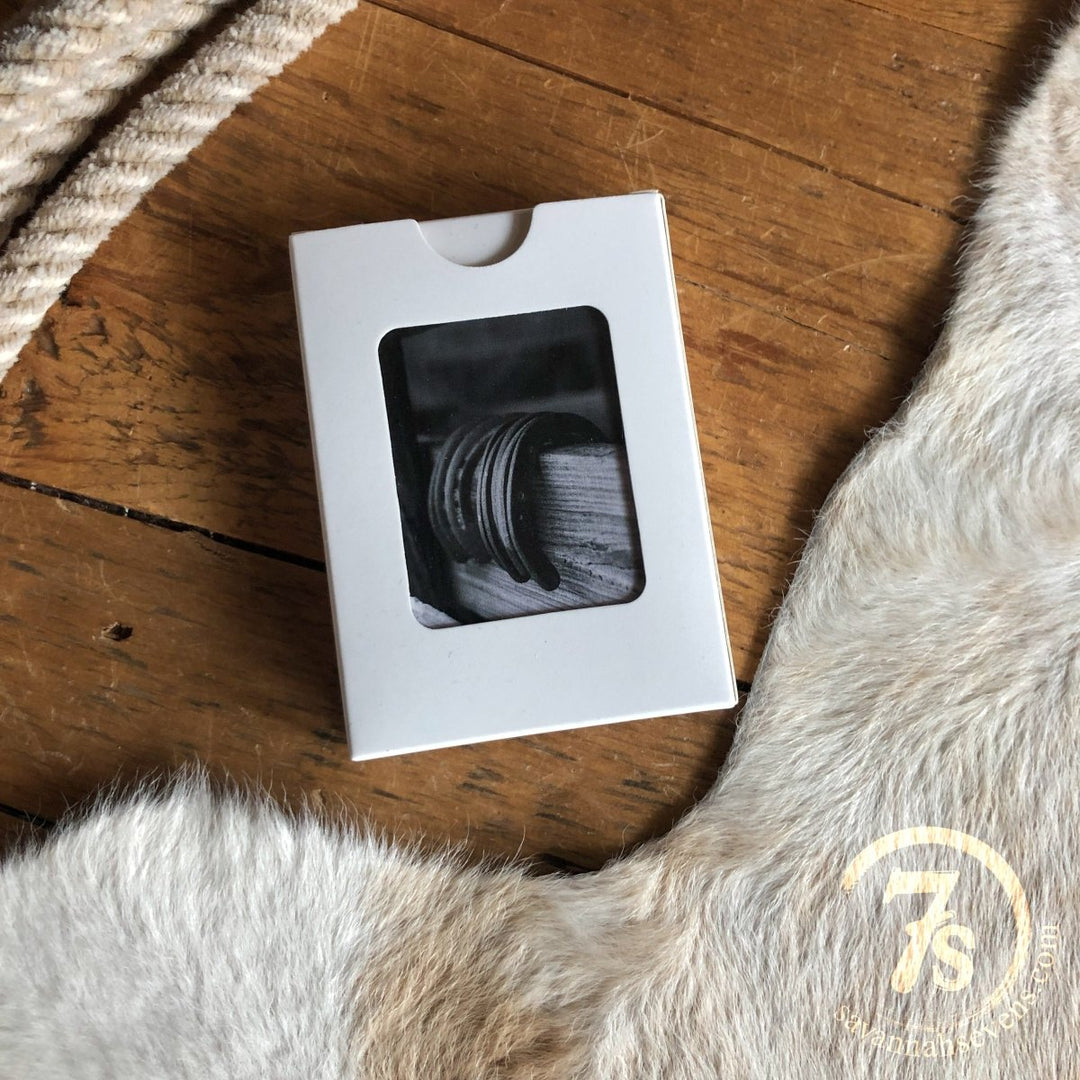 A small white box with a transparent window displays Savannah Sevens western life{&}style Lucky Playing Cards on a wooden floor beside a woven item and light rug, with western card deck motifs and a lucky horseshoe adding charm.