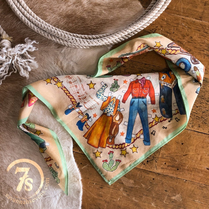 The Paper Dolls Neck Scarf by Savannah Sevens, featuring hand-drawn Western motifs—cowboy boots, hats, shirts, jeans, cacti, horseshoes, and stars—lies folded on a wooden floor near a rope. savannahsevens.com is visible.