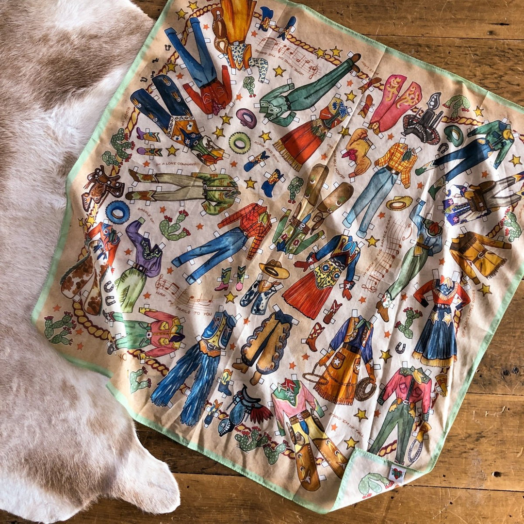 The Paper Dolls Neck Scarf by Savannah Sevens, featuring hand-drawn western-themed clothing, boots, hats, horses, and accessories, is laid flat on a wooden floor beside a cowhide rug.
