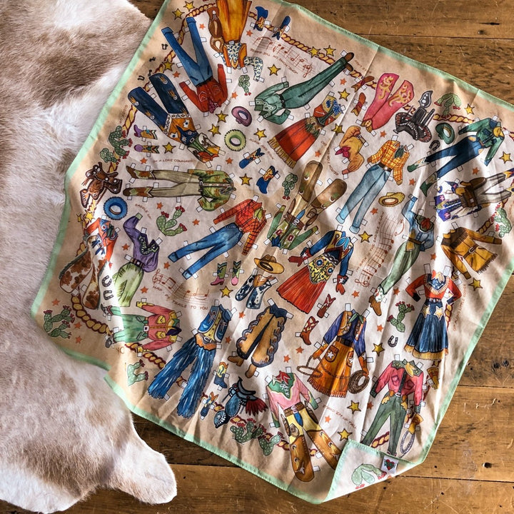The Paper Dolls Neck Scarf by Savannah Sevens, featuring hand-drawn western-themed clothing, boots, hats, horses, and accessories, is laid flat on a wooden floor beside a cowhide rug.