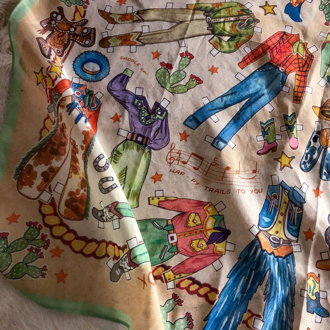 The Paper Dolls Neck Scarf by Savannah Sevens features hand-drawn cowboy outfits, hats, boots, cacti, and musical notes with western stars. "Happy Trails To You" appears throughout this colorful vintage accessory.