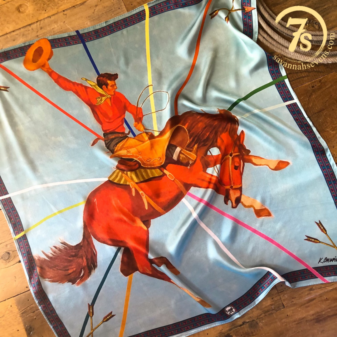 The Rodeo King Wild Rag by Savannah Sevens western life{&}style is a vibrant silk scarf featuring a rodeo cowboy on a bucking horse, a blue background with multicolored lines, aztec trim, and the 7S logo in the top right corner.