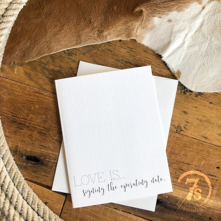 The Hungry Canyon "Signing The Operating Note Card," made from white linen paper and featuring the phrase “LOVE IS... signing the operating note,” rests on a wooden surface with a white envelope, cowhide rug, and rope in the background.