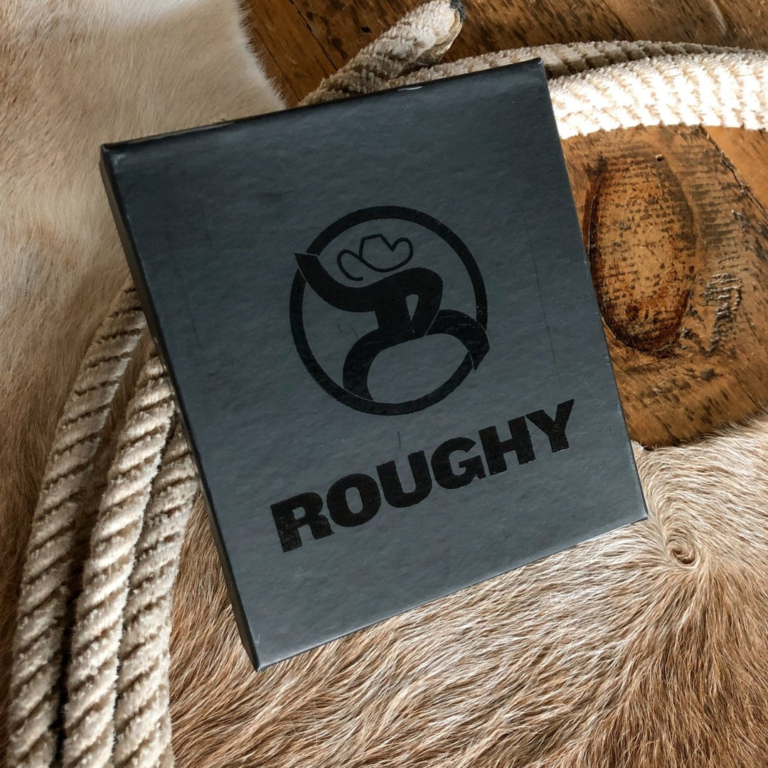 A black square box with "ROUGHY" and a stylized logo on the lid sits on coiled rope atop cowhide and wood, pairing perfectly with the Storden Money Clip from Savannah Sevens western life{&}style for added style. By Hooey. 
