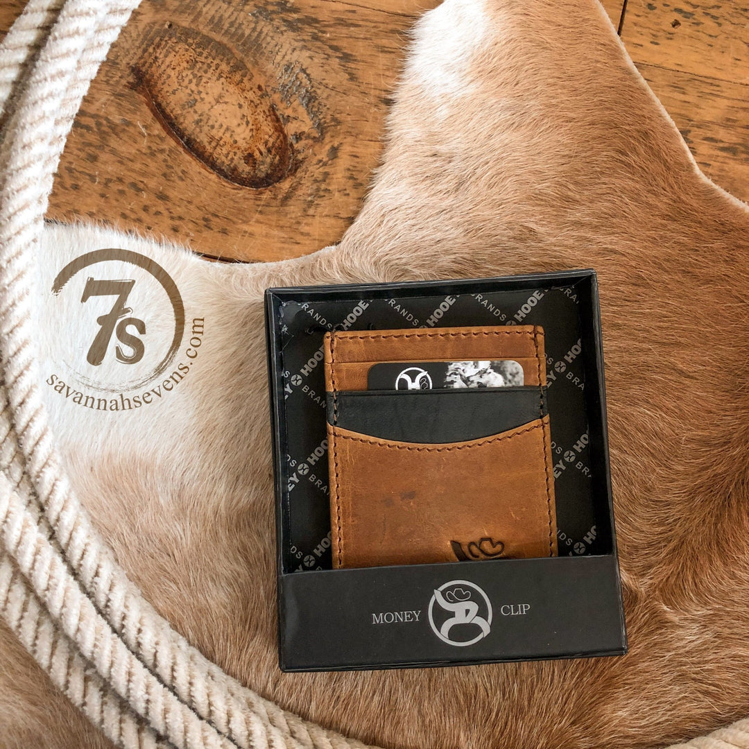 The Storden Money Clip by Savannah Sevens western life{&}style, in distressed brown leather and a black gift box, sits on a tan cowhide rug beside a coiled rope and stamped 7s savannahsevens.com logo.
