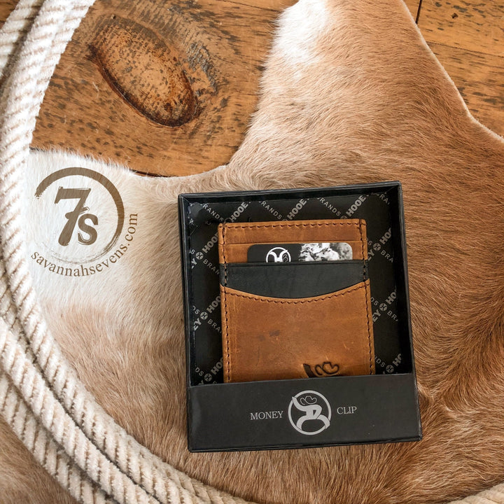 The Storden Money Clip by Savannah Sevens western life{&}style, in distressed brown leather and a black gift box, sits on a tan cowhide rug beside a coiled rope and stamped 7s savannahsevens.com logo.