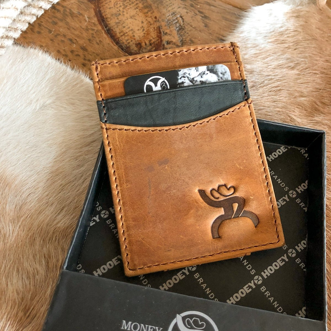 The Storden Money Clip by Savannah Sevens western life{&}style, a brown distressed leather card holder with the brand's logo, holds two cards and sits on a black branded box placed on cowhide. By Hooey. 