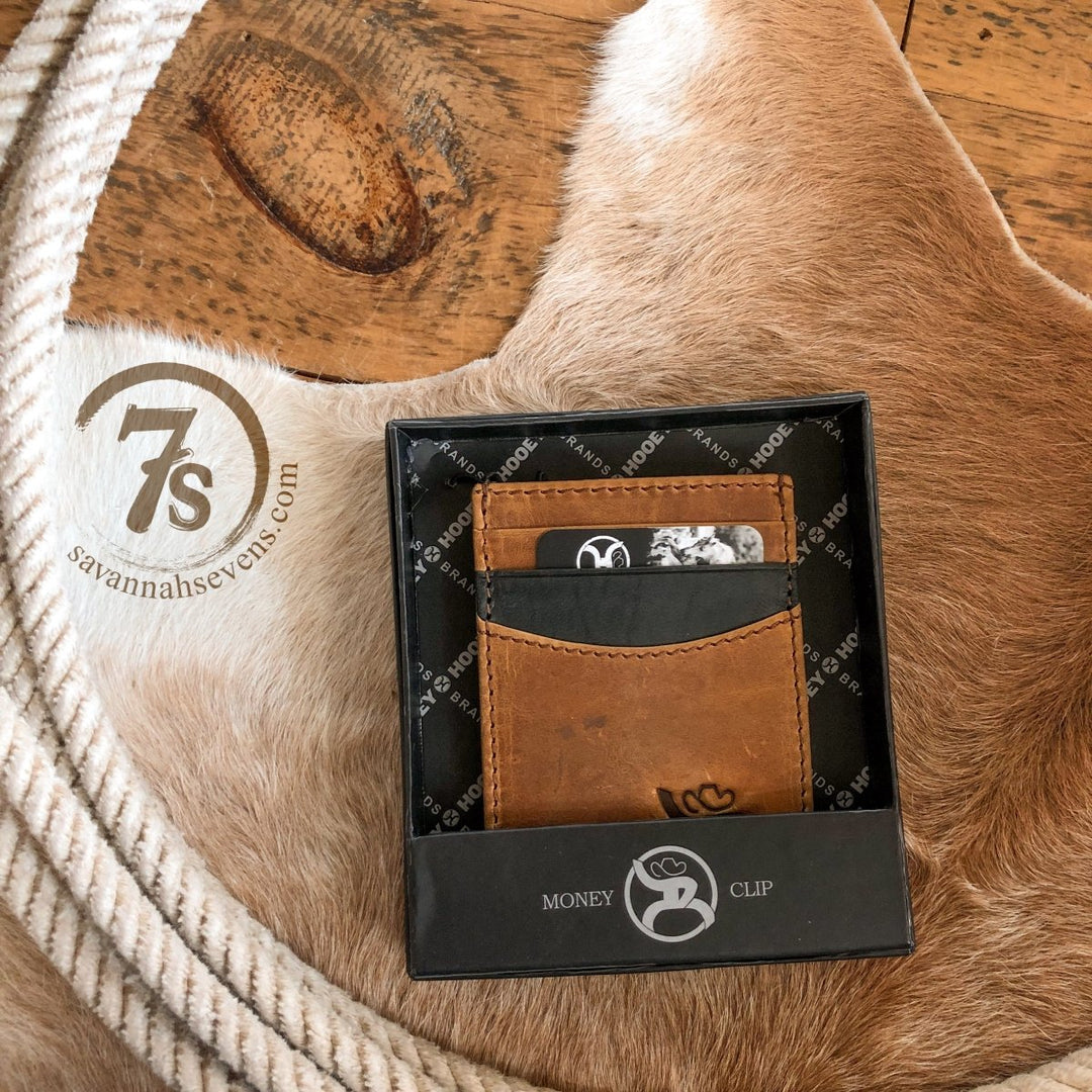 The Storden Money Clip by Savannah Sevens western life{&}style, in distressed brown leather and a black gift box, sits on a tan cowhide rug beside a coiled rope and stamped 7s savannahsevens.com logo.