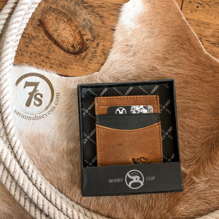 The Storden Money Clip by Savannah Sevens western life{&}style, in distressed brown leather and a black gift box, sits on a tan cowhide rug beside a coiled rope and stamped 7s savannahsevens.com logo.