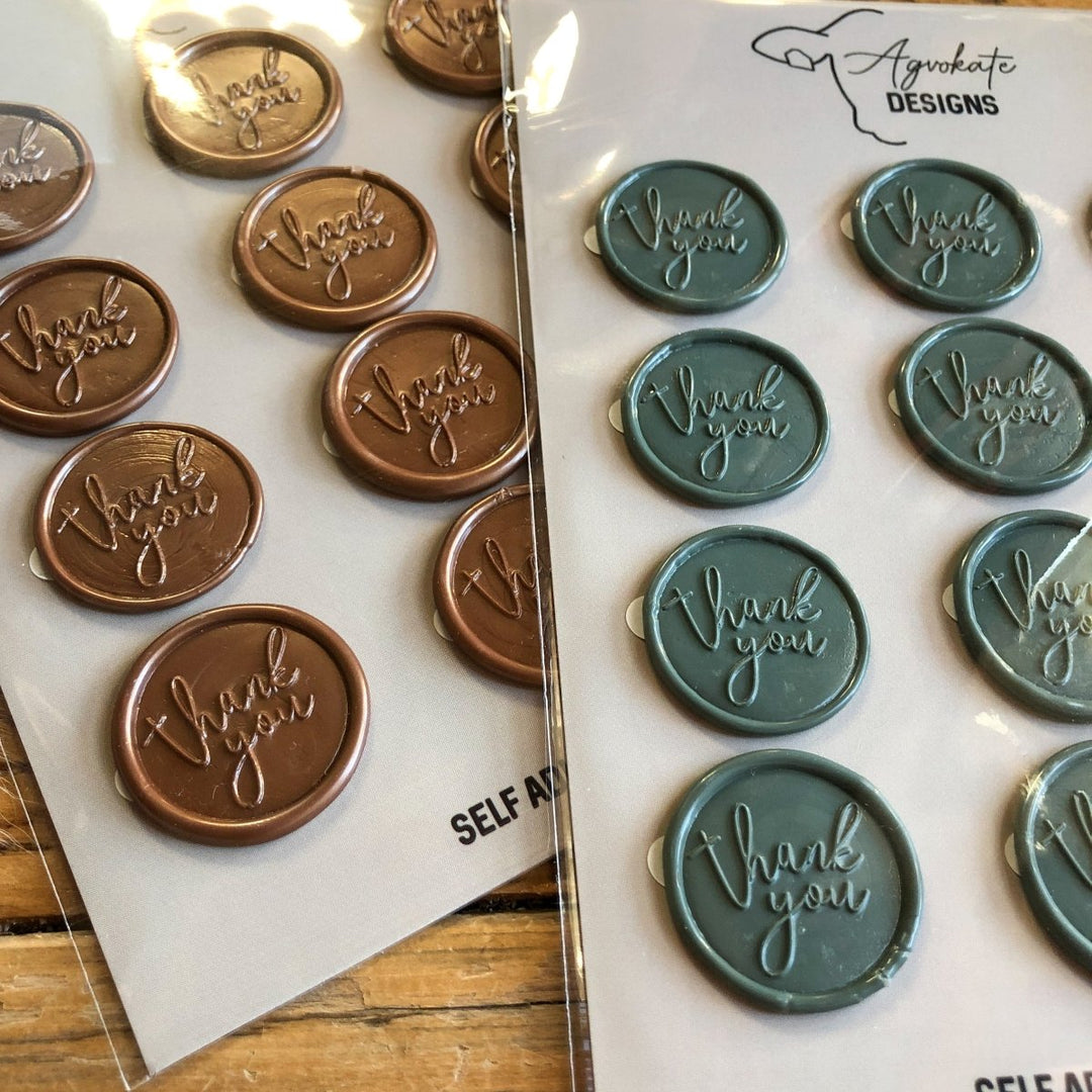 Two self-adhesive Thank You Wax Seal stickers by Savannah Sevens western life{&}style—one brown, one teal—each embossed with “thank you” in cursive, rest on a wooden surface.