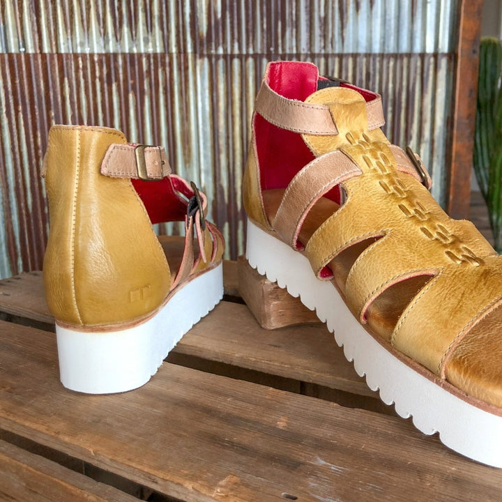 A pair of The Cotulla mustard dip dye leather platform sandals by Savannah Sevens western life{&}style, featuring thick white soles and red lining, displayed on rustic wood with a corrugated metal background.
