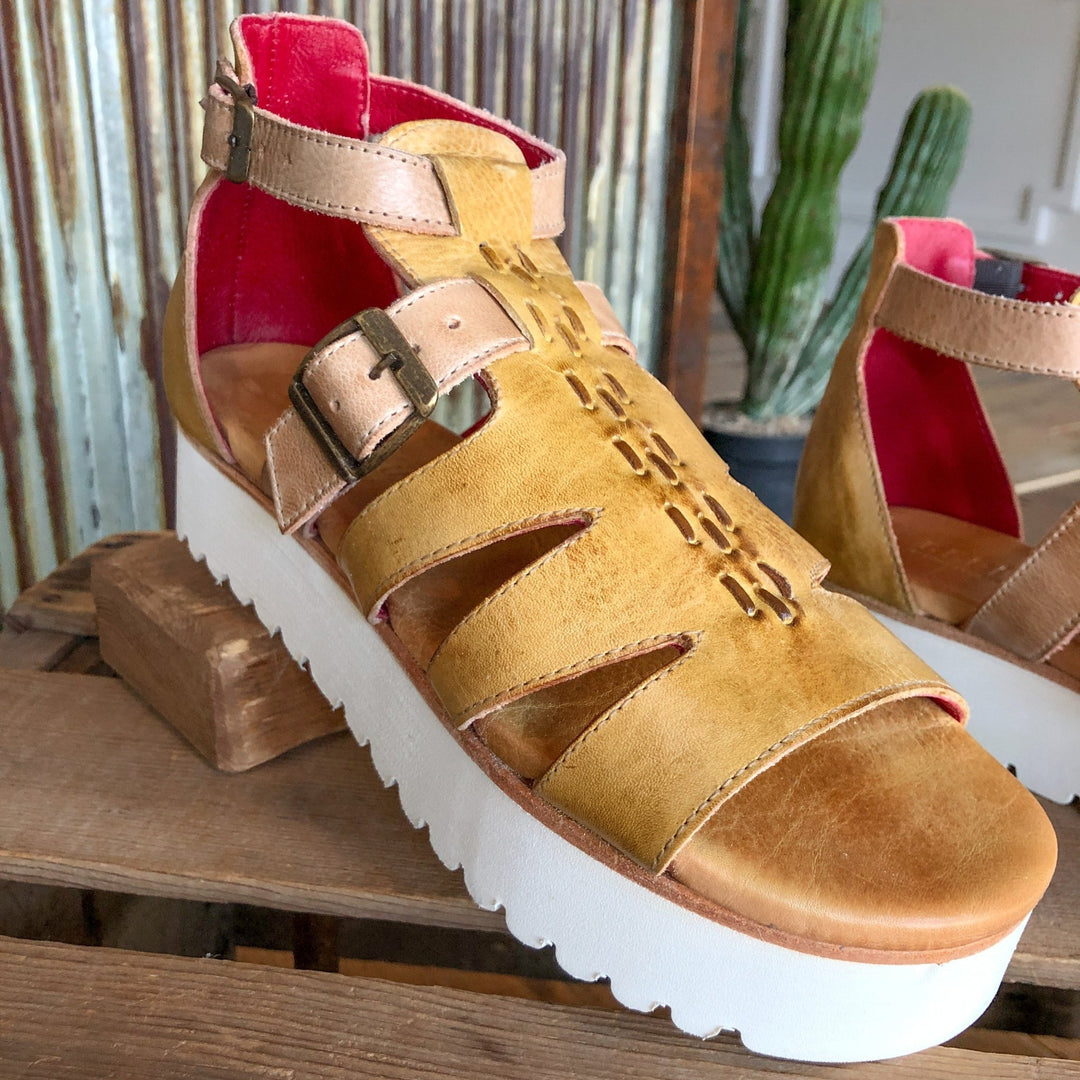 A close-up of The Cotulla leather platform sandal by Savannah Sevens western life{&}style in tan with a thick white sole, buckle strap, geometric cutouts, and bright pink lining, shown on wood with a cactus background.