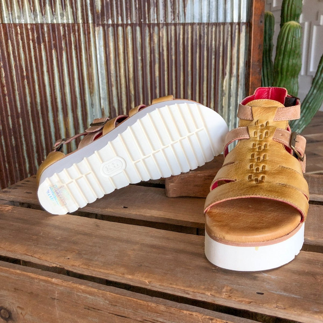 The Cotulla platform sandals by Savannah Sevens western life{&}style, featuring mustard dip-dyed leather and thick white soles, are shown on rustic wood with one upright and the other sideways, set before a corrugated metal wall and cactus. Sandals made by BedStu. 