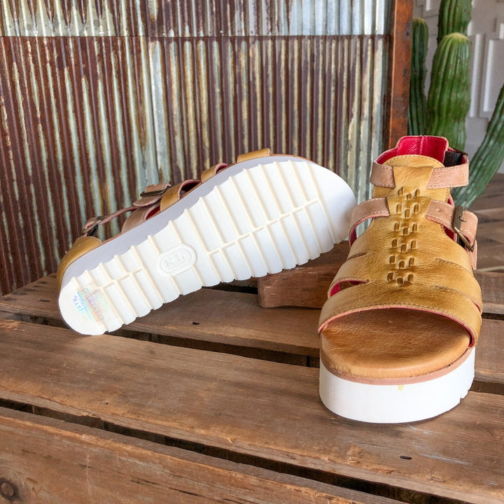 The Cotulla platform sandals by Savannah Sevens western life{&}style, featuring mustard dip-dyed leather and thick white soles, are shown on rustic wood with one upright and the other sideways, set before a corrugated metal wall and cactus. Sandals made by BedStu. 