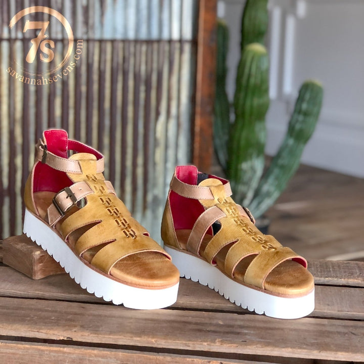 The Cotulla platform sandals by Savannah Sevens western life{&}style feature mustard dip dye leather, white zigzag soles, and red lining, shown on rustic wood with a cactus and corrugated metal wall in the background.