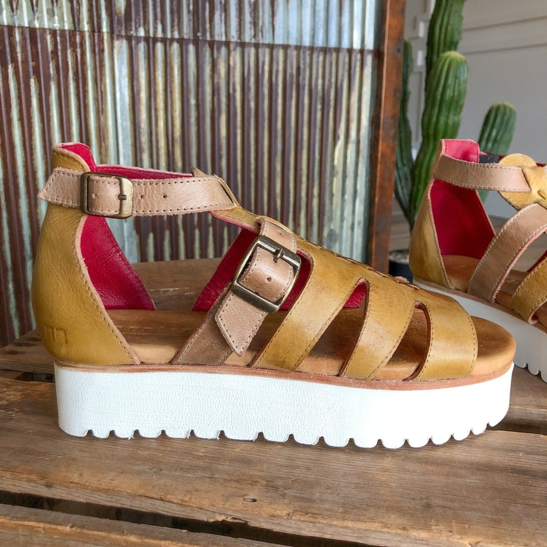 The Cotulla by Savannah Sevens western life{&}style is a leather platform sandal with multiple straps, two buckles, a thick white sole, tan and mustard dip-dye leather, and red interior lining, shown on wood with cactus and corrugated metal backdrop.