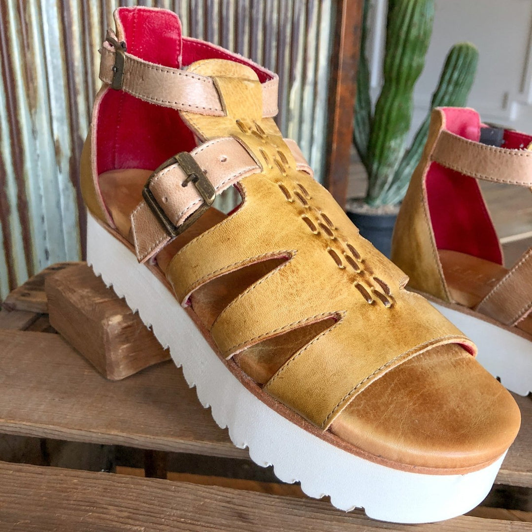 A close-up of The Cotulla leather platform sandal by Savannah Sevens western life{&}style in tan with a thick white sole, buckle strap, geometric cutouts, and bright pink lining, shown on wood with a cactus background.