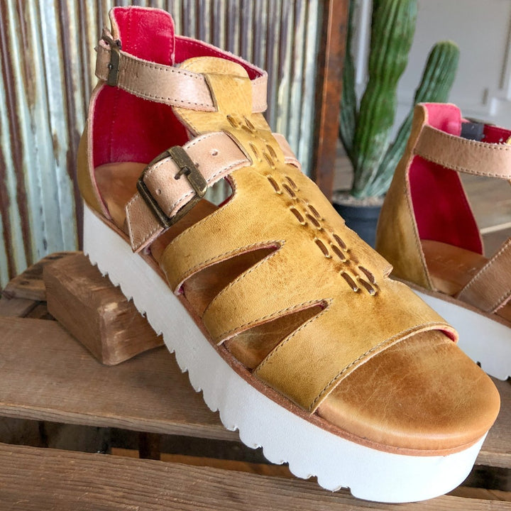 A close-up of The Cotulla leather platform sandal by Savannah Sevens western life{&}style in tan with a thick white sole, buckle strap, geometric cutouts, and bright pink lining, shown on wood with a cactus background.