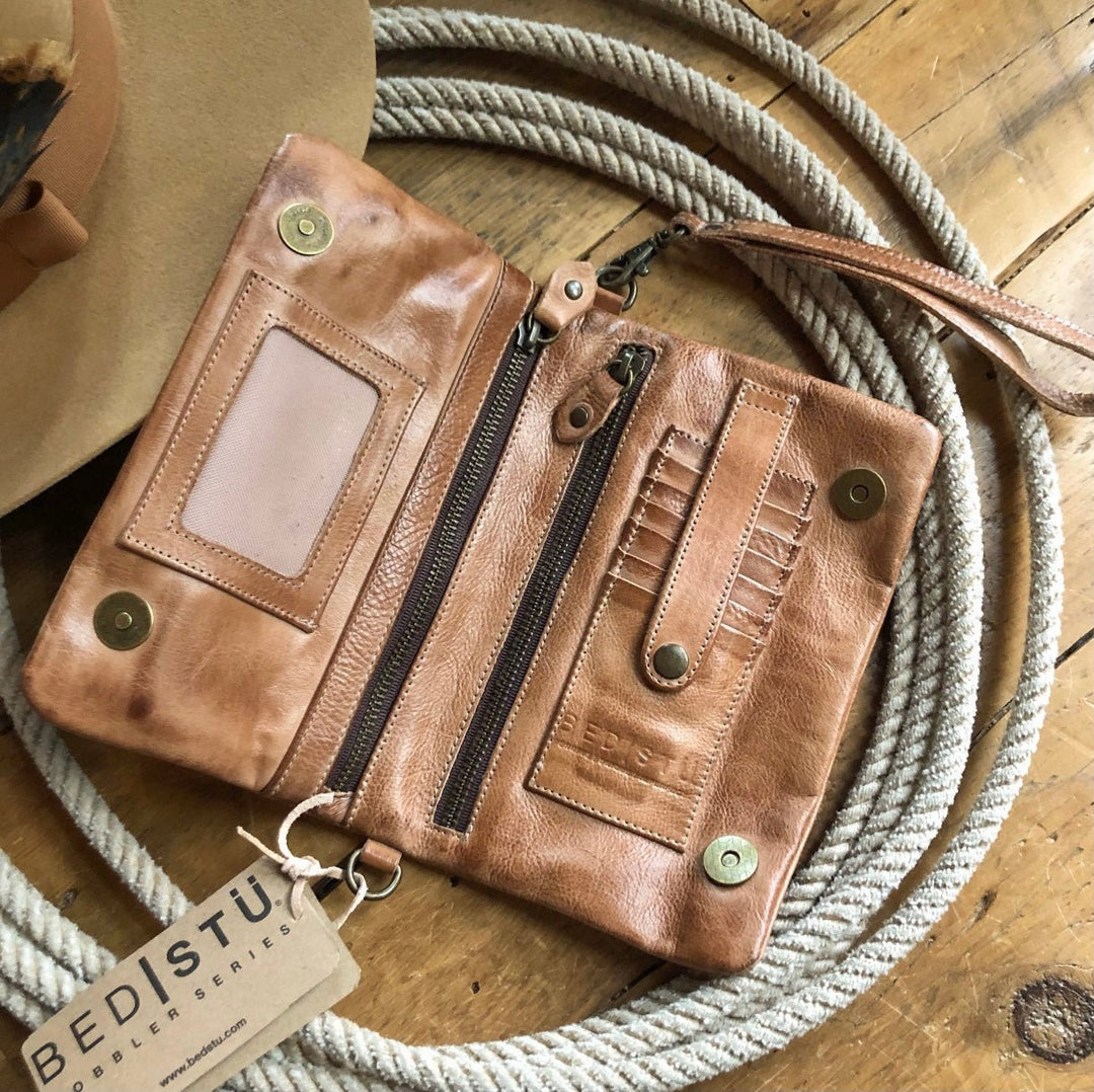 The Roca wallet by Savannah Sevens western life{&}style, featuring a tan dip-dye leather finish, card slots, zipper pocket, and snap closures, sits open on wood near a rope. A Bed|stü tag and part of a hat are also visible. By BedStu. 
