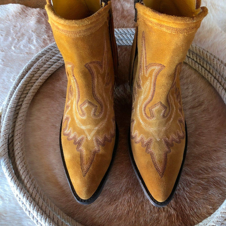 The Tejas by Savannah Sevens western life{&}style: yellow suede western boots with decorative stitching, displayed on a round cowhide and bordered by subtle leather fringe accents.